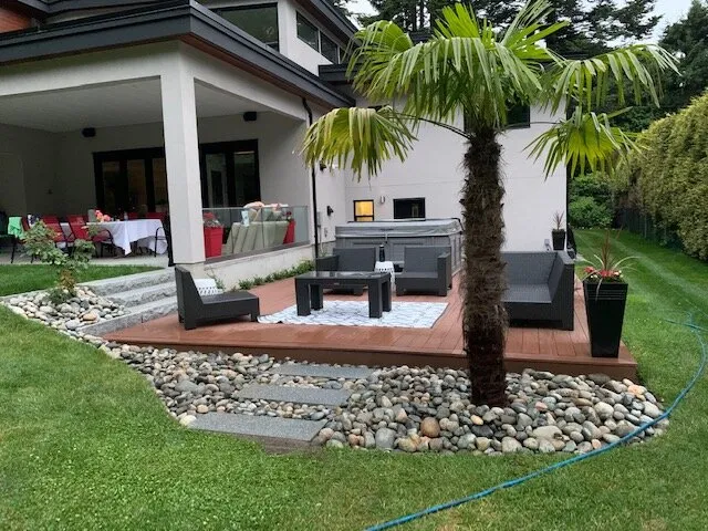Covered back patio with sunken lounge deck, river-rock landscaping, potted palm, and hot tub.