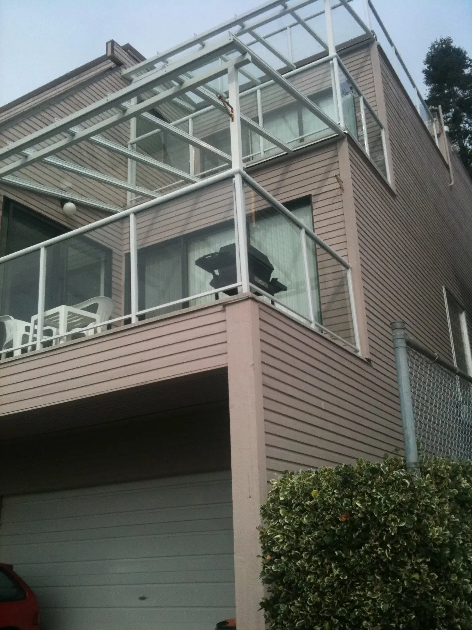 Low-angle view of the worn vinyl siding, glass deck railing, and plastic patio furniture below.
