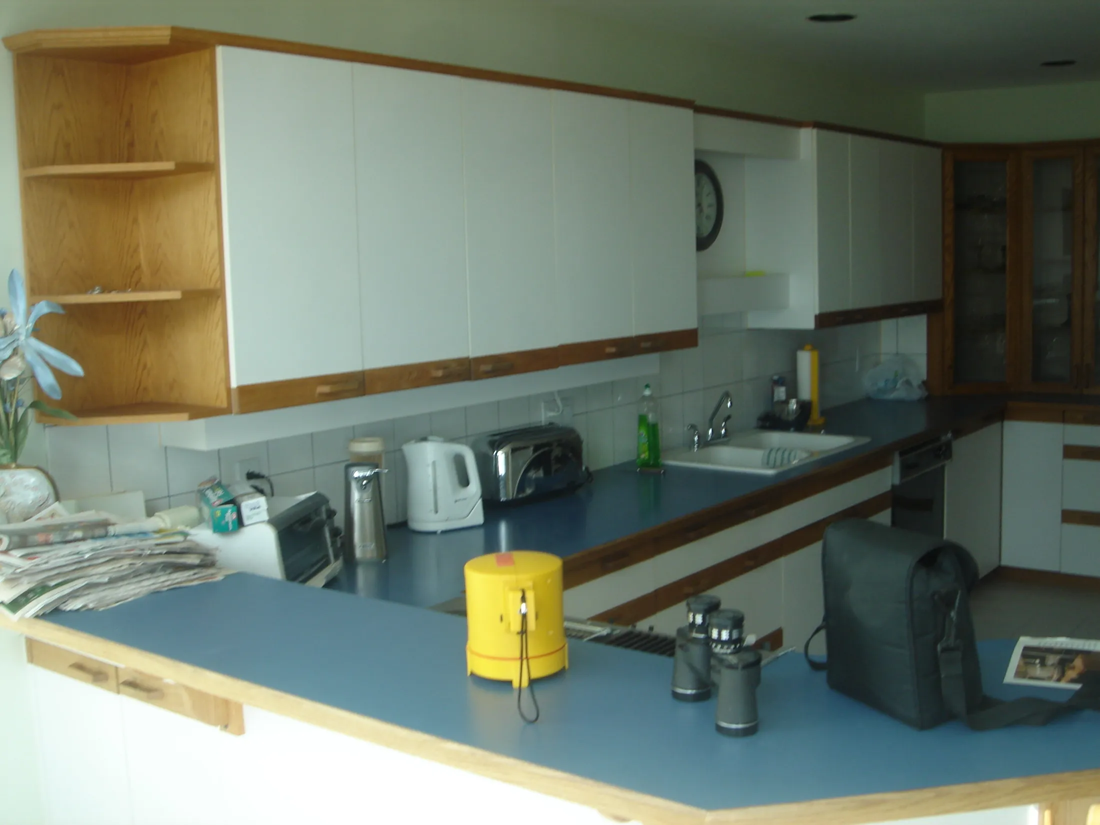 Original kitchen with dated blue laminate counters, oak-trimmed white cabinets, and a built-in wall clock.