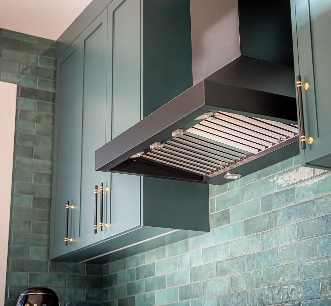 Angled close-up of range hood over sage-green cabinets with brass pulls and green subway tile.