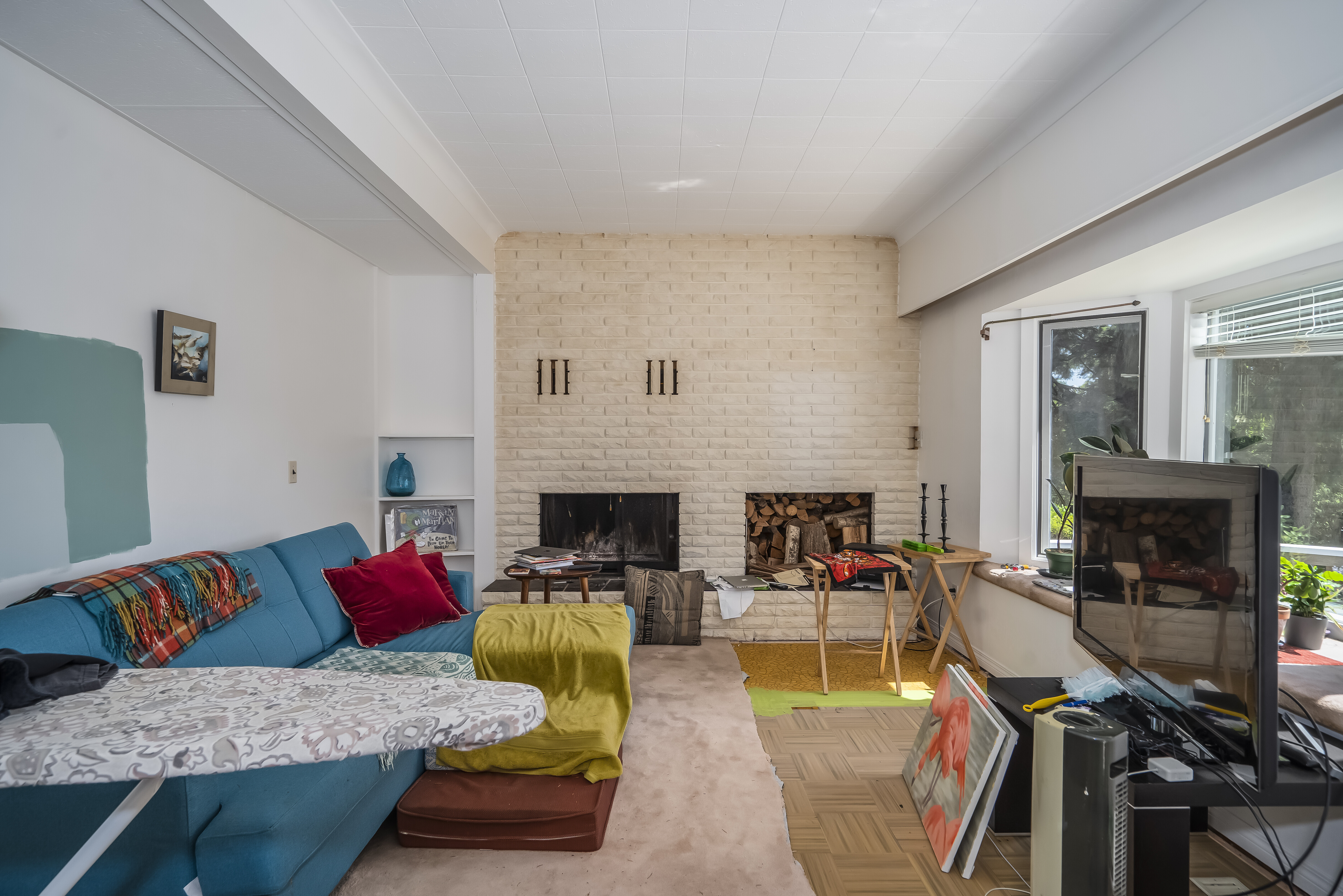 Pre-renovation living room with painted brick fireplace, dated tile ceiling, parquet flooring, and cluttered furnishings.