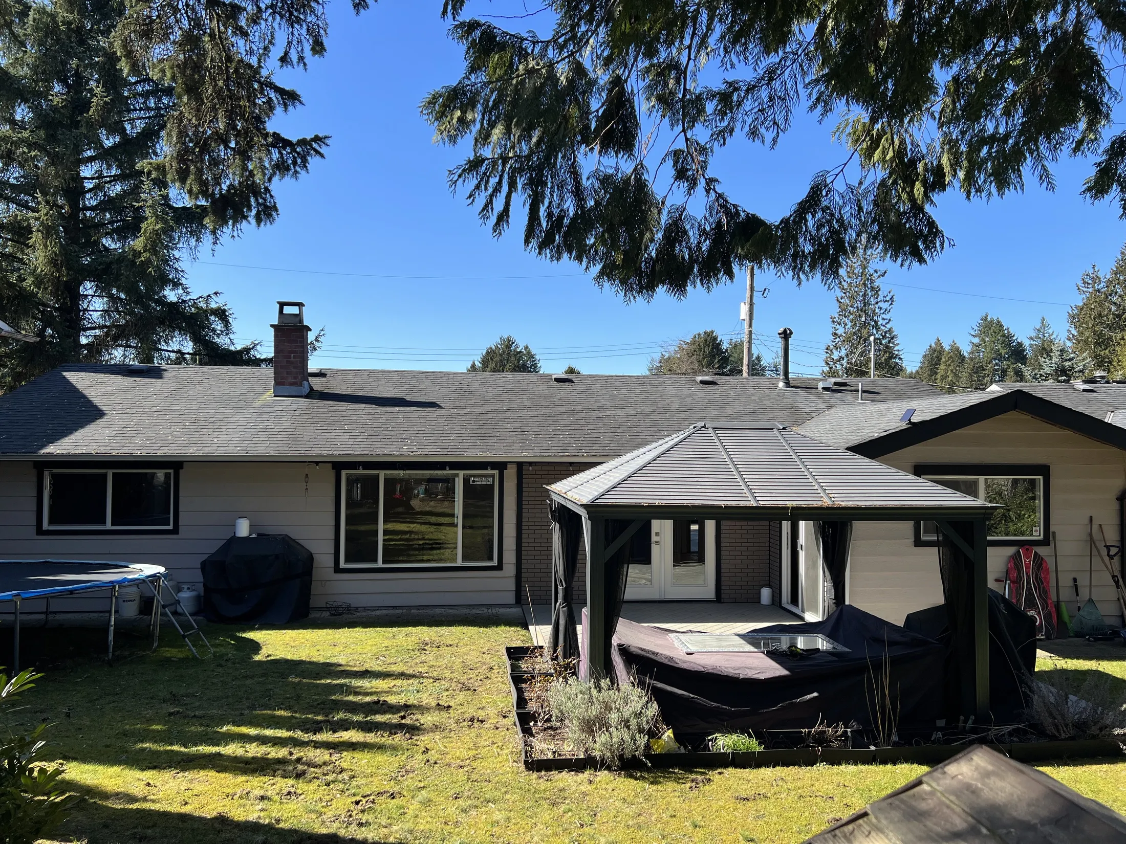 Backyard before renovation, showing the dated ranch exterior, brick chimney, trampoline, and covered gazebo.