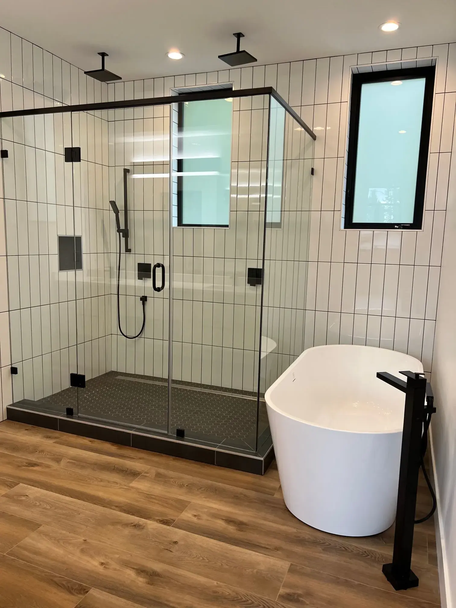 Primary bath with glass-enclosed rain shower, matte black fixtures, freestanding tub, and vertical subway tile.