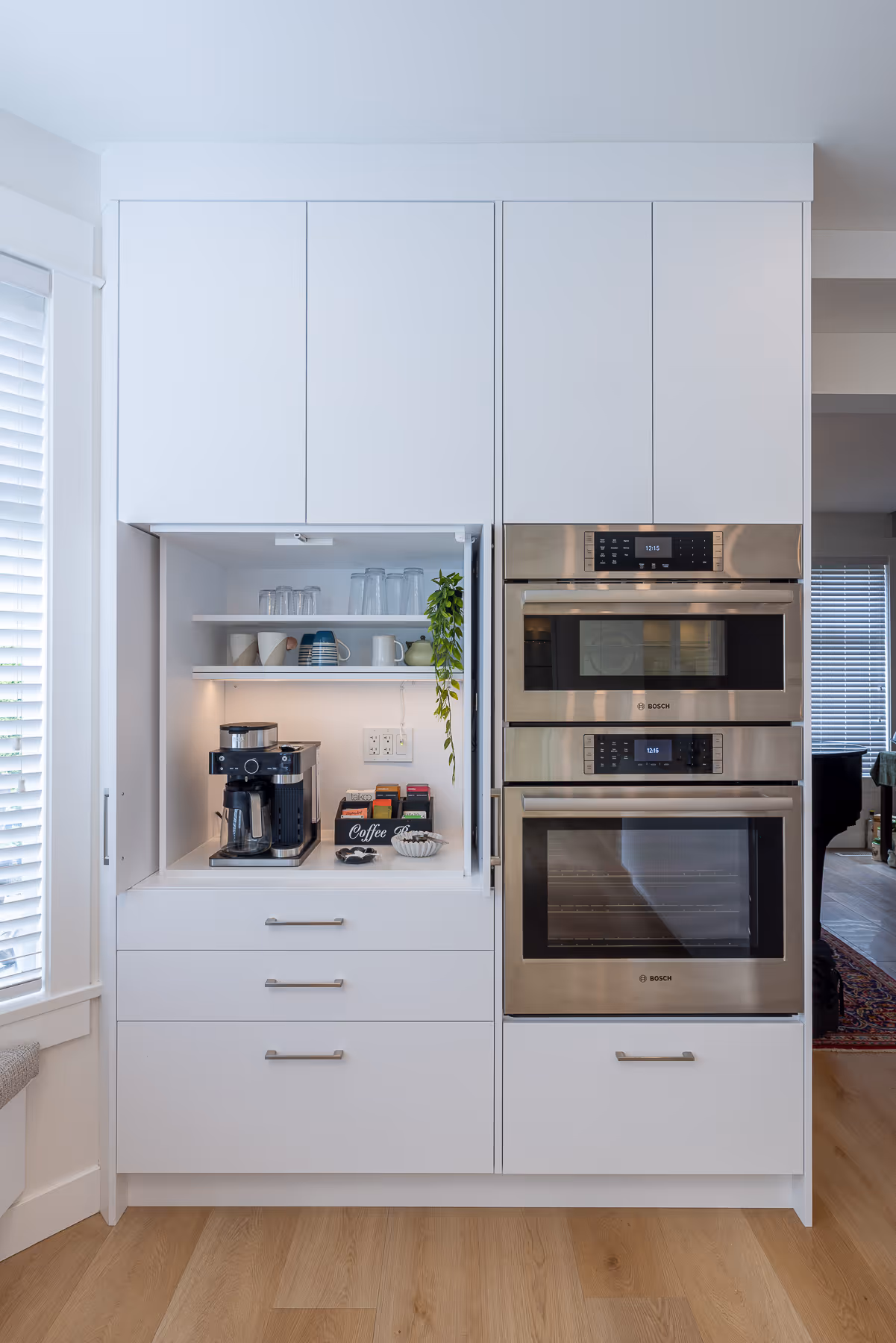 Open coffee station cabinet reveals glass shelving beside stainless double wall ovens.