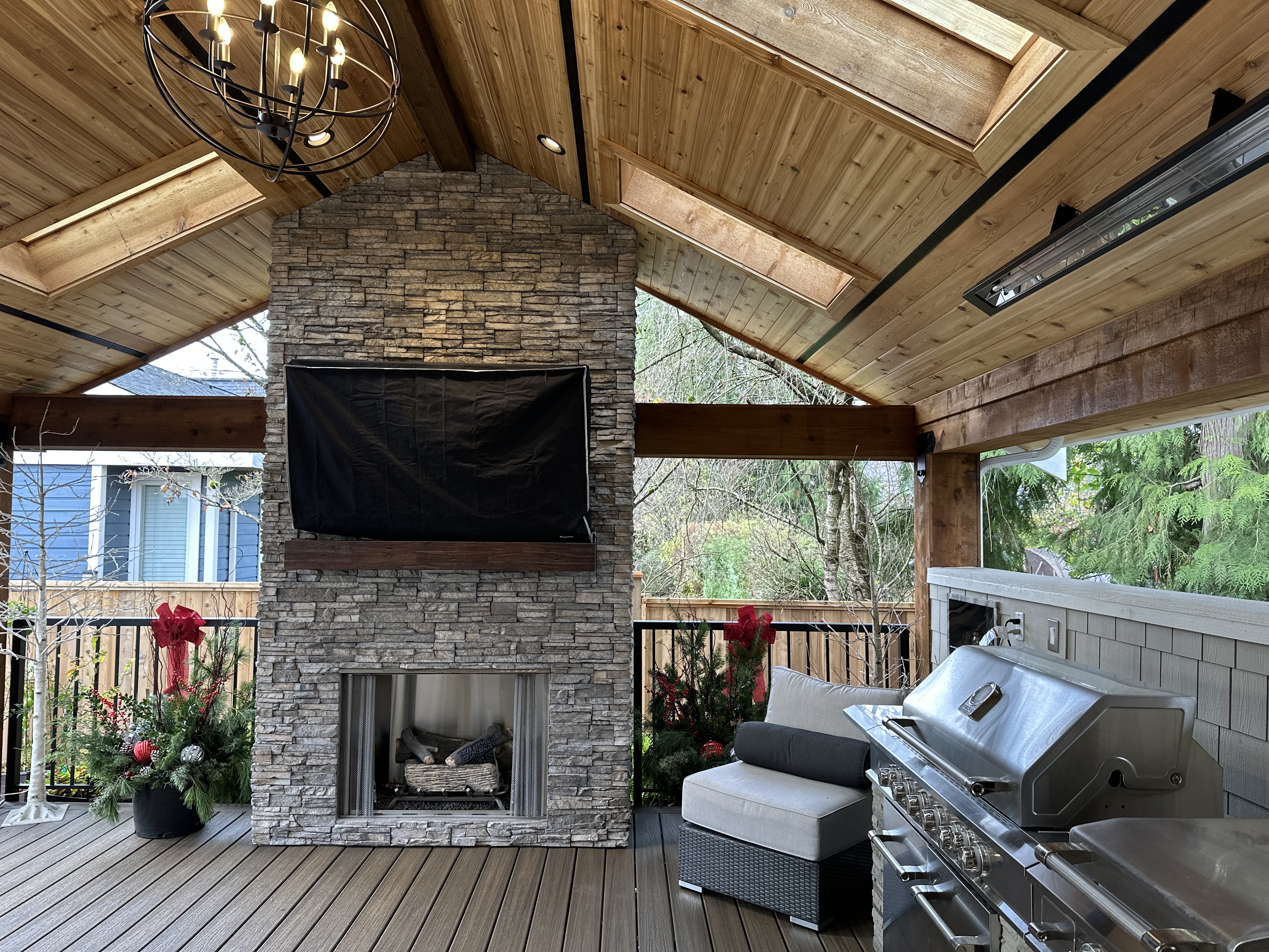 Covered patio with stone fireplace, mounted TV, chandelier, and stainless grill station, decorated for the holidays.