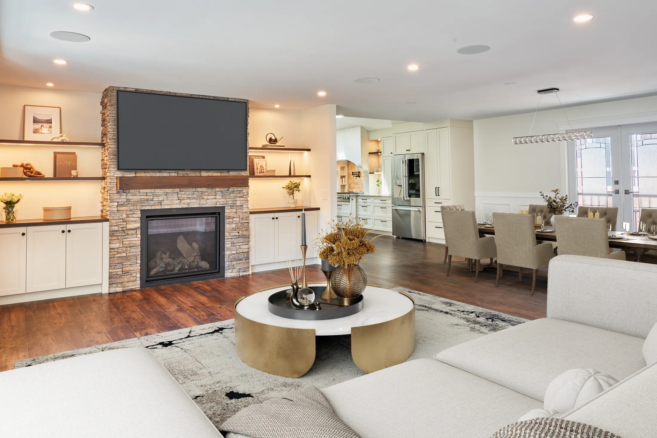 Living room with a stacked-stone fireplace, mounted TV, and round brass coffee table, open to the kitchen.