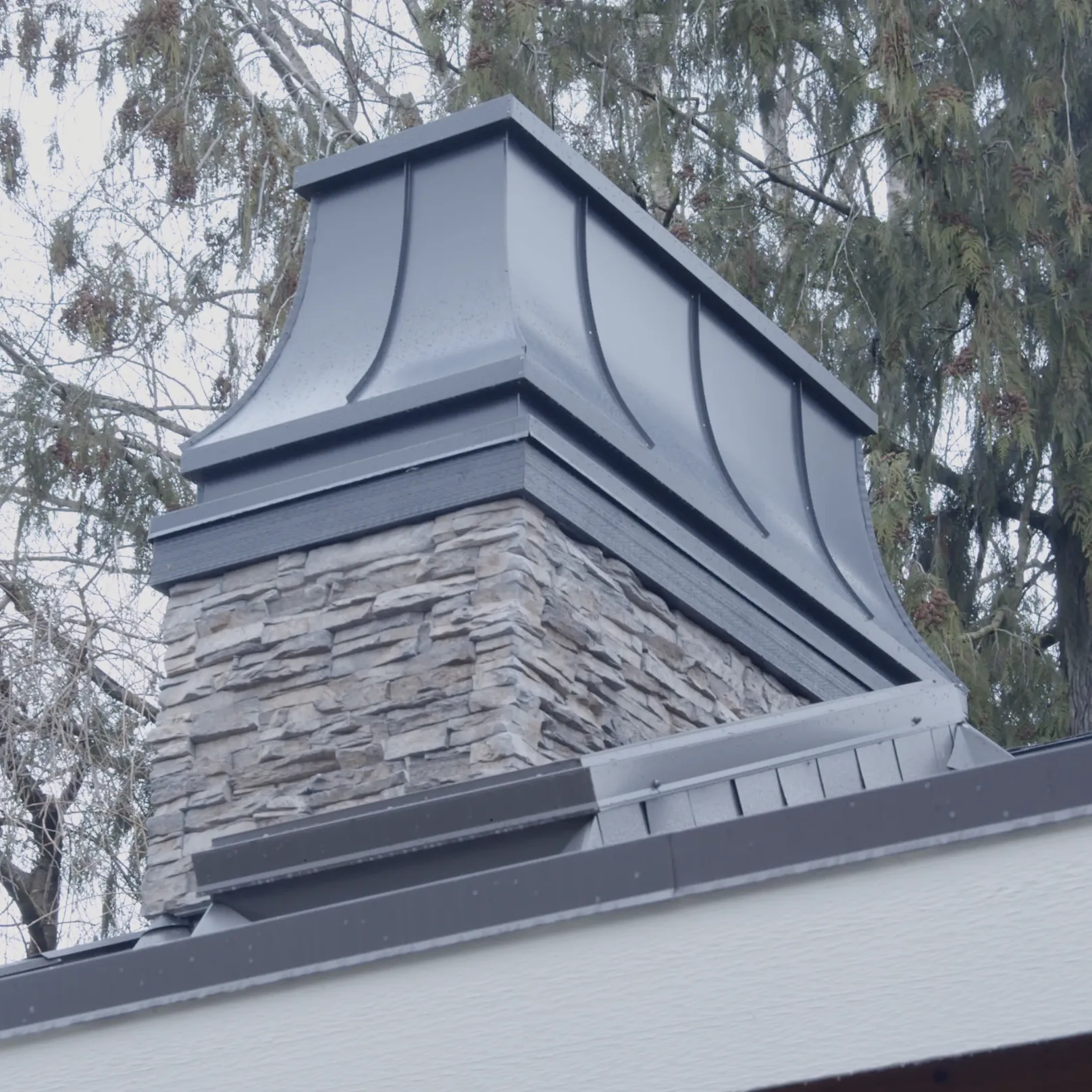 Low-angle view of the stacked-stone chimney and its dark metal cap against the tree canopy.