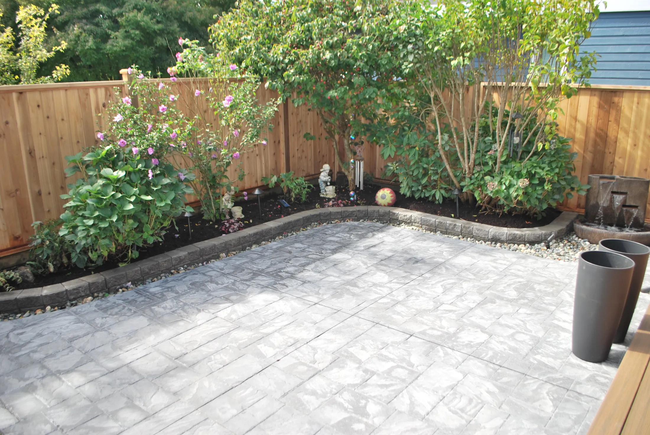 Backyard patio corner with stamped concrete, flowering shrubs, and a tiered stone water fountain along the fence.