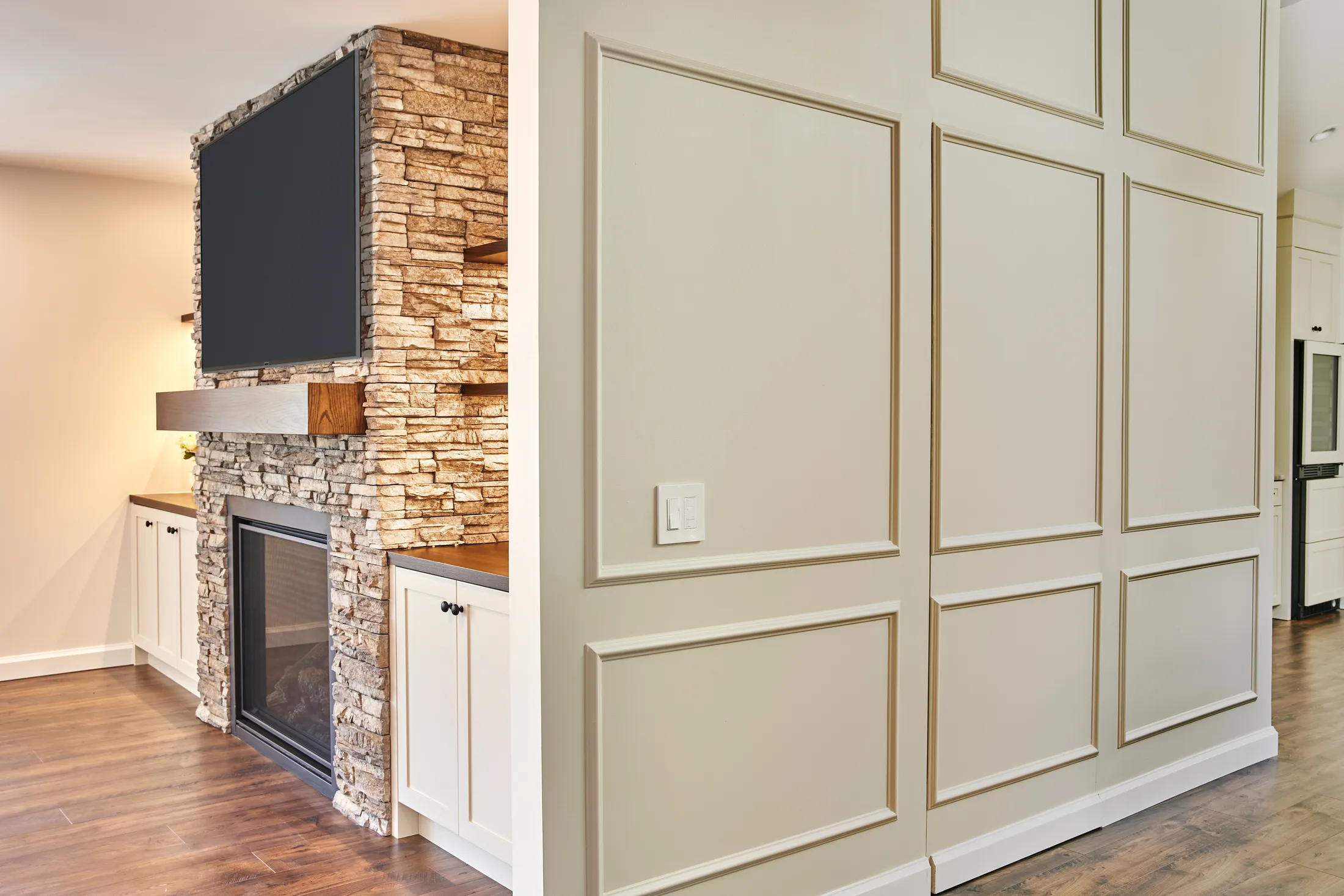 Stacked-stone fireplace with mounted TV beside a paneled wainscoted wall opening to the kitchen hallway.