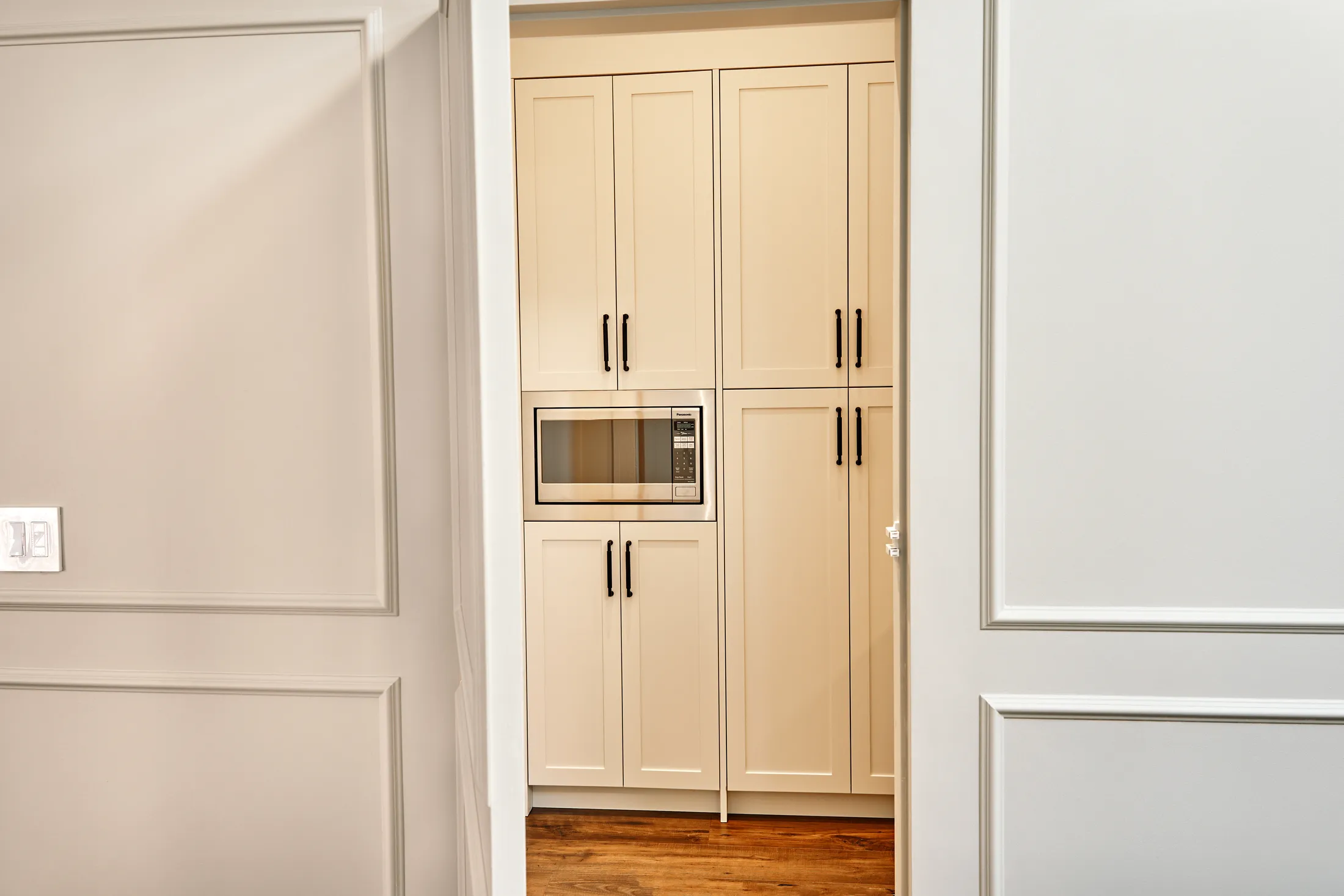 Tall pantry cabinetry with a built-in microwave and black hardware, viewed through the hallway doorway.