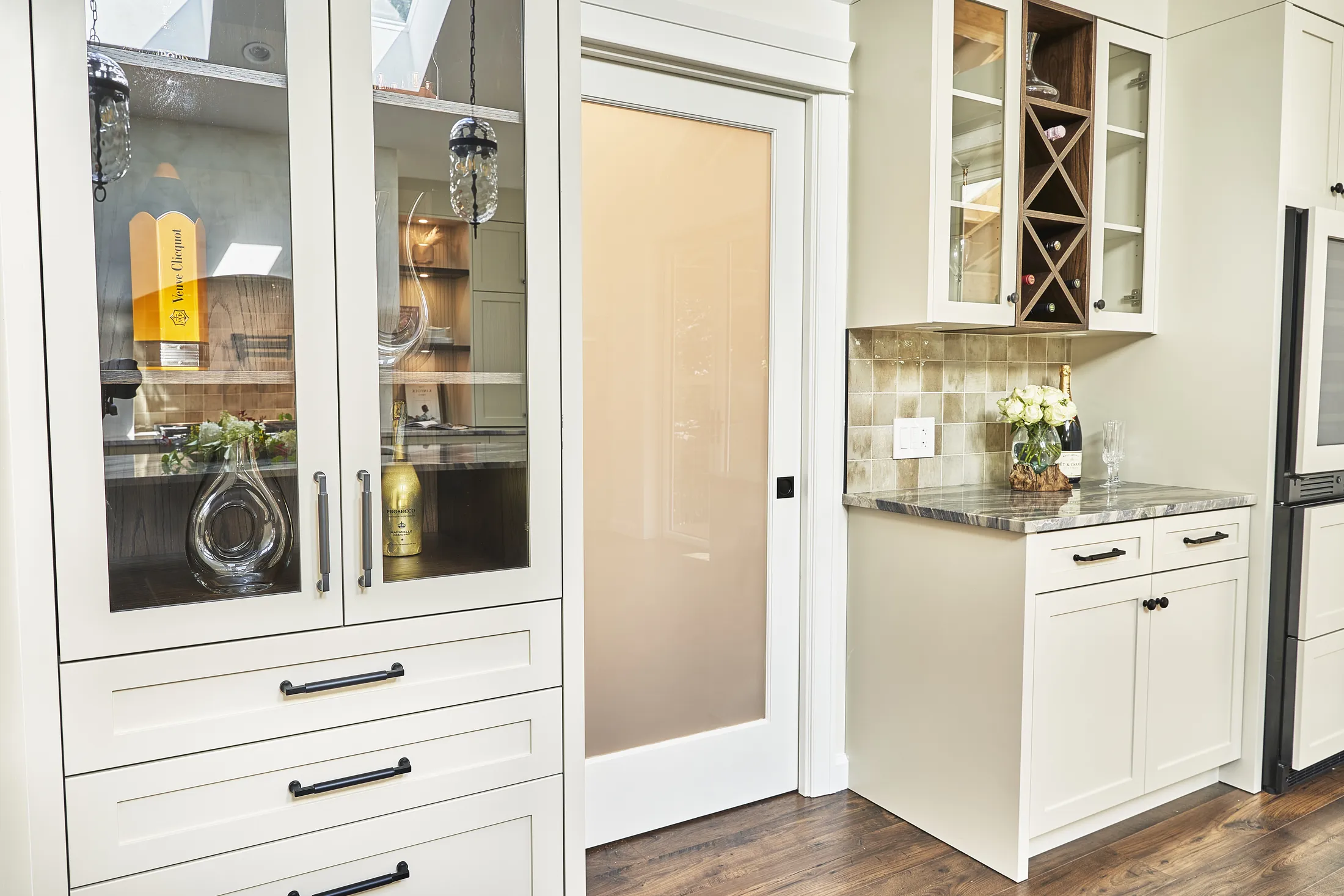 Glass-front bar cabinet with wine bottles and glassware beside a frosted-glass pocket door and wine cubby.