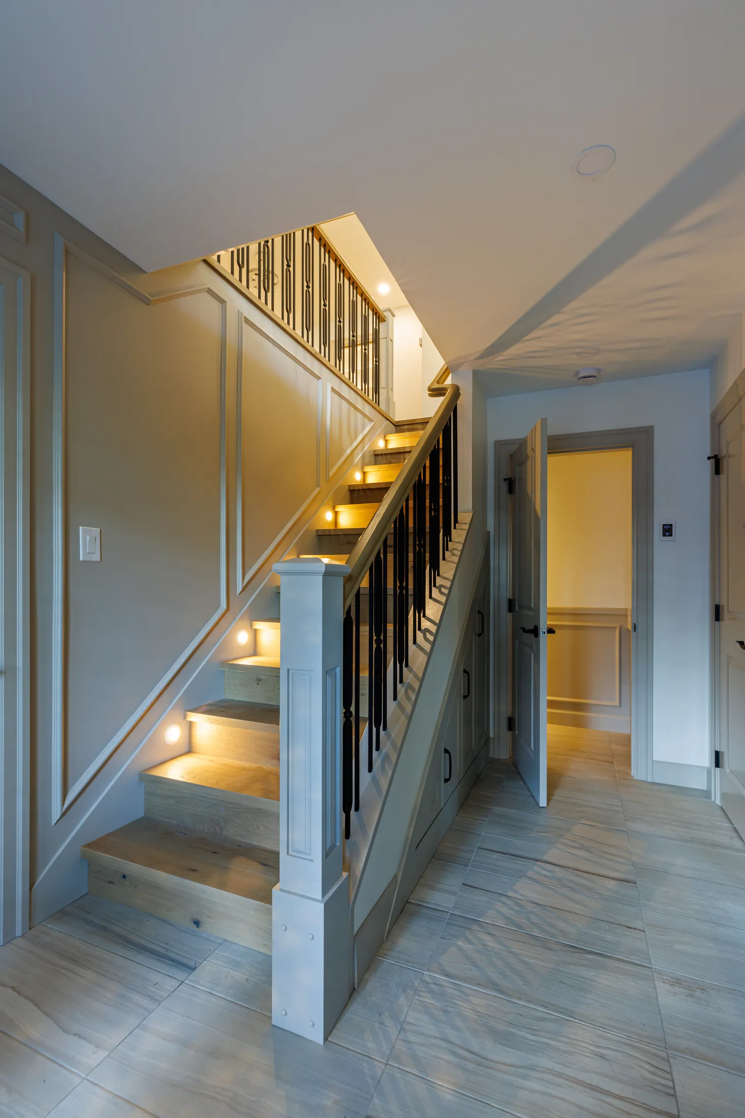 Wood staircase with black wrought-iron balusters and step lighting descending to a tiled lower hallway.