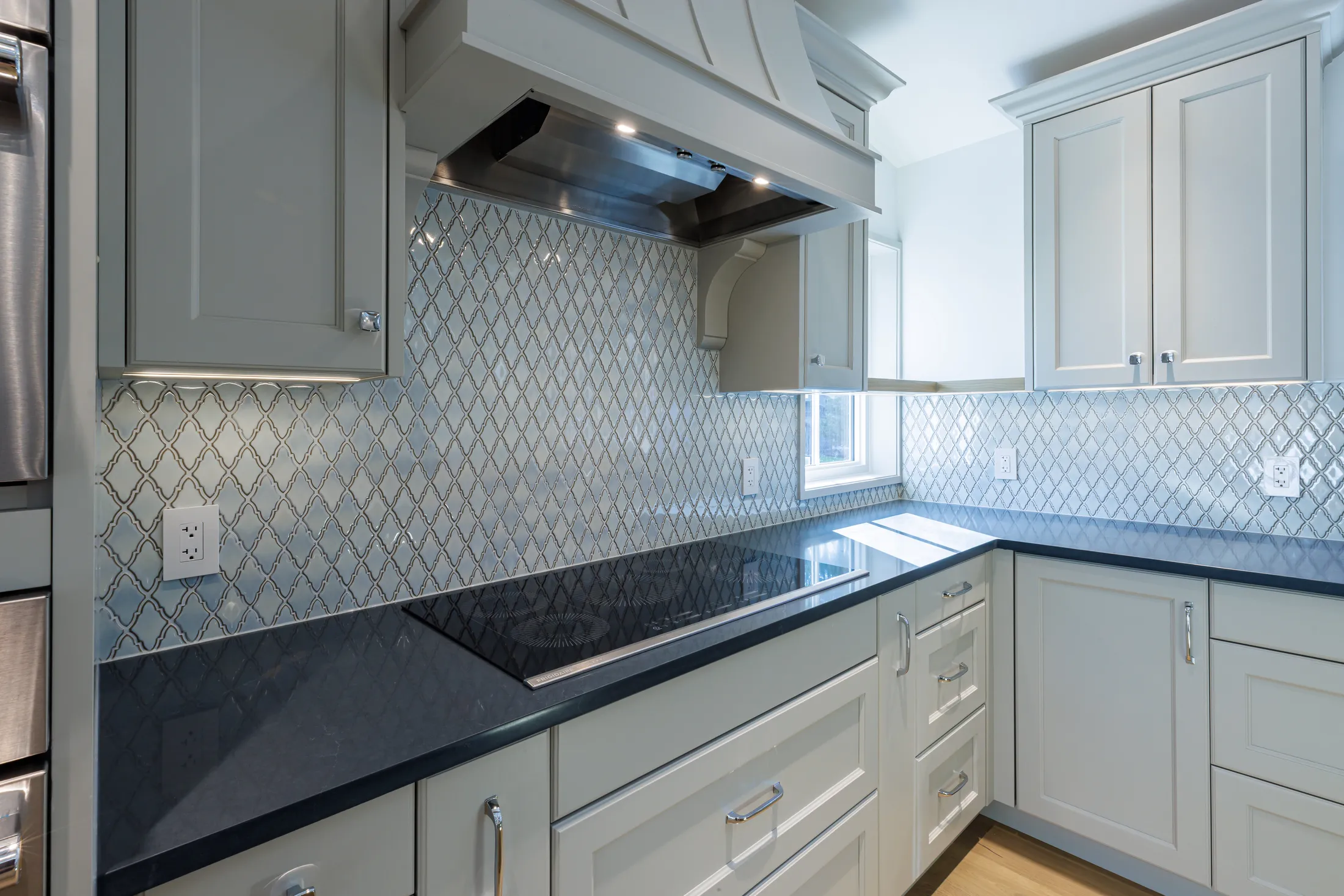 Corner view of the tiled cooktop backsplash wrapping toward a window, with black stone counters.