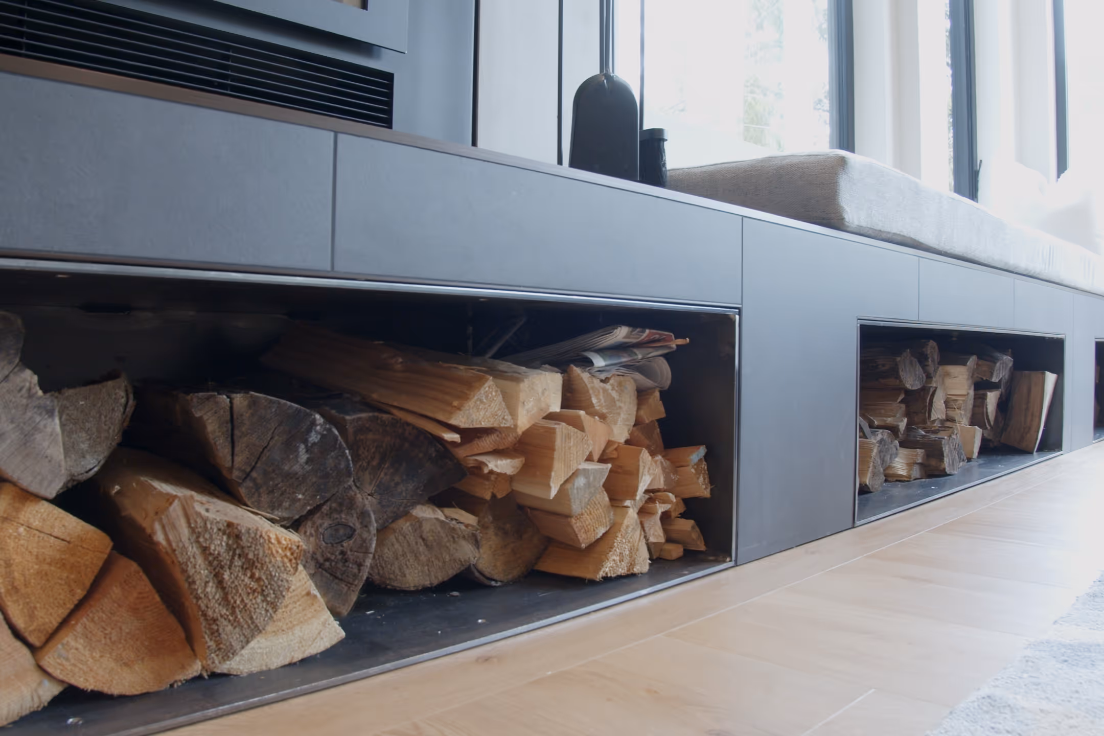 Built-in firewood storage niche below a window seat, split cedar logs stacked on oak flooring.