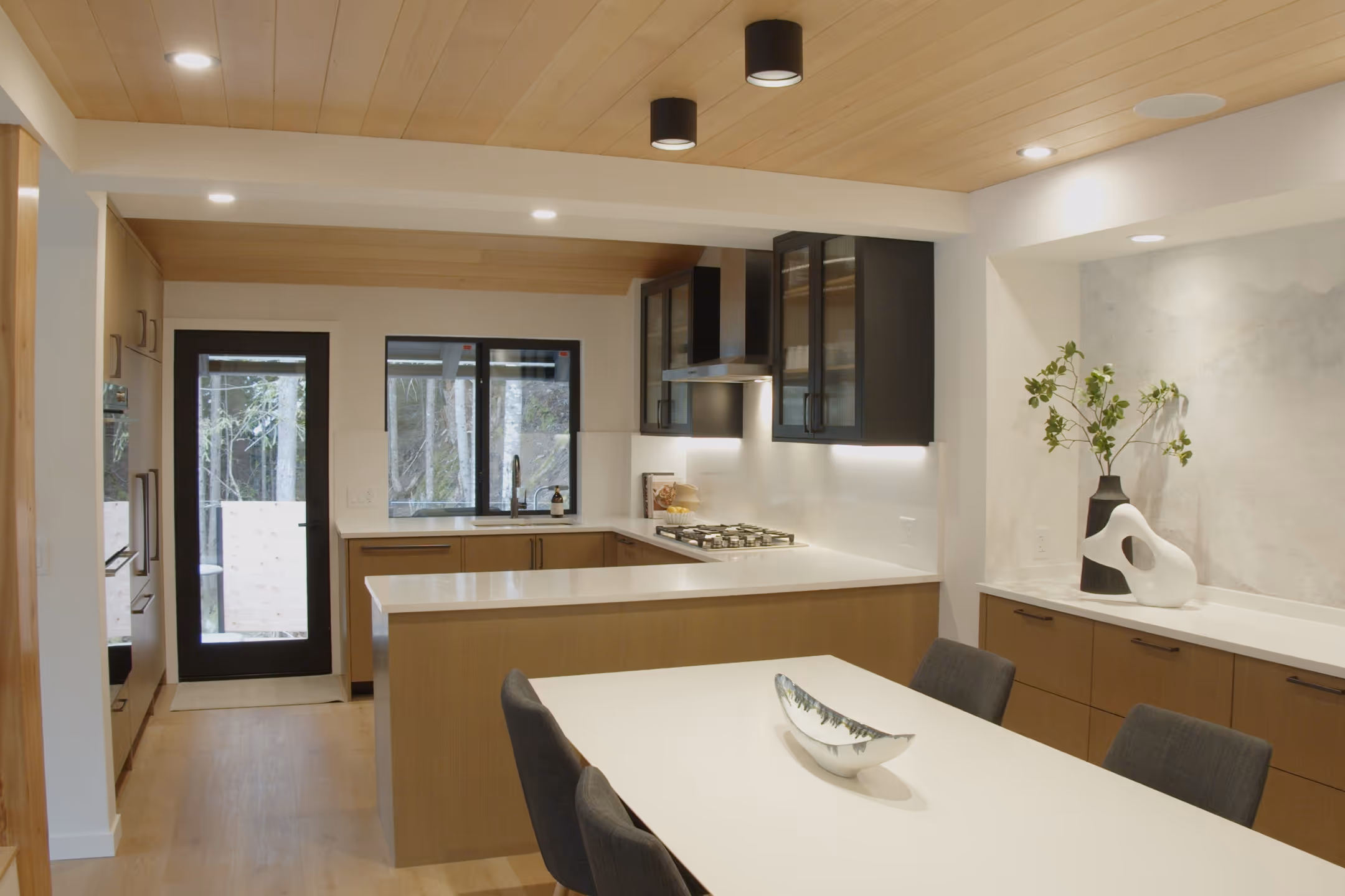 Open kitchen and dining area with cedar-plank ceiling, black glass-front cabinetry, white quartz counters, walnut table.