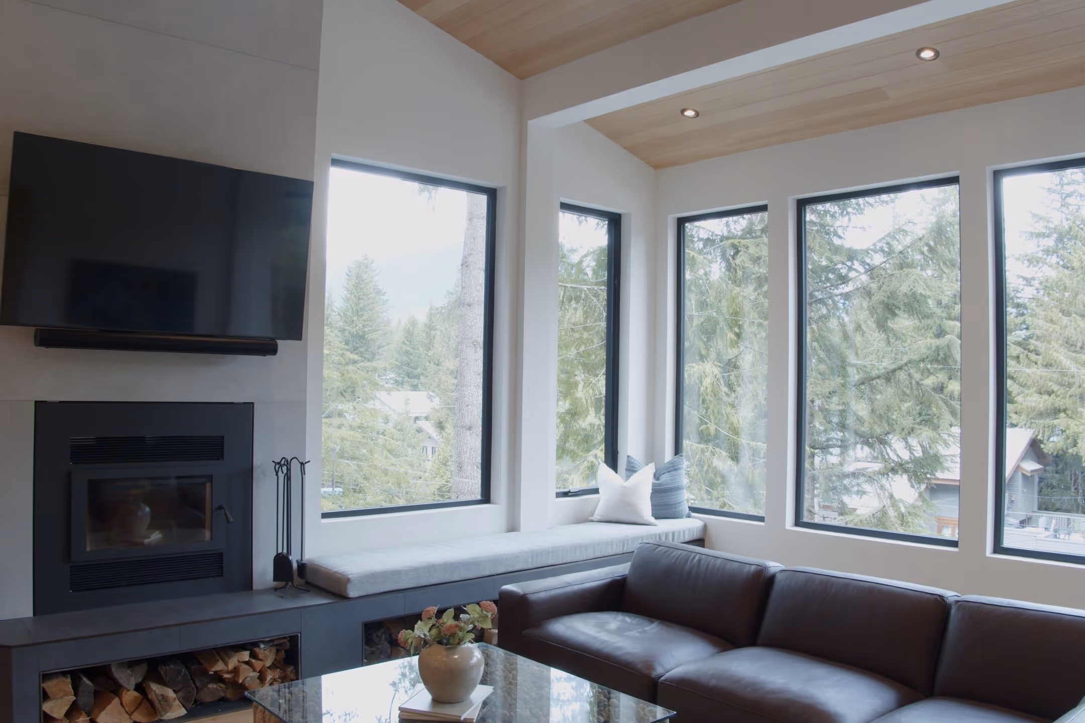 Living room with vaulted wood ceiling, wall-mounted TV over a fireplace, built-in window seat facing forest.