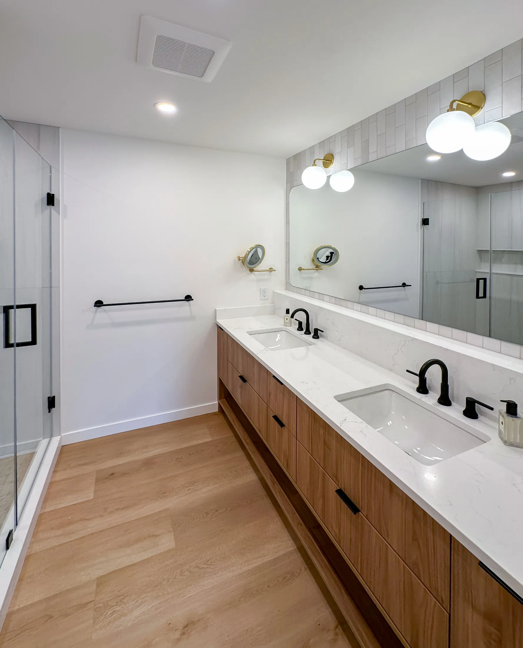 Primary bathroom double vanity: black wood cabinetry, white quartz counter, matte-black fixtures, brass sconces, large mirror.