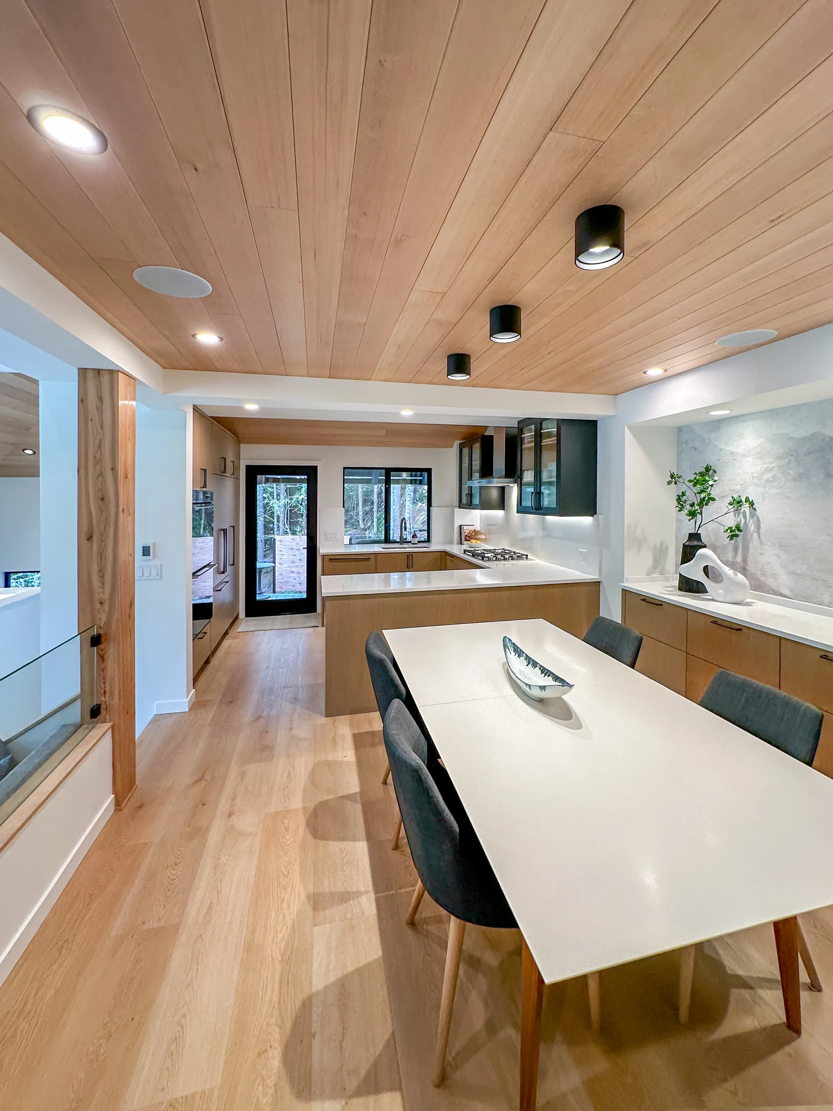 Kitchen viewed toward the entry hall, black cylindrical pendant lights, glass-front cabinets, wood dining table.