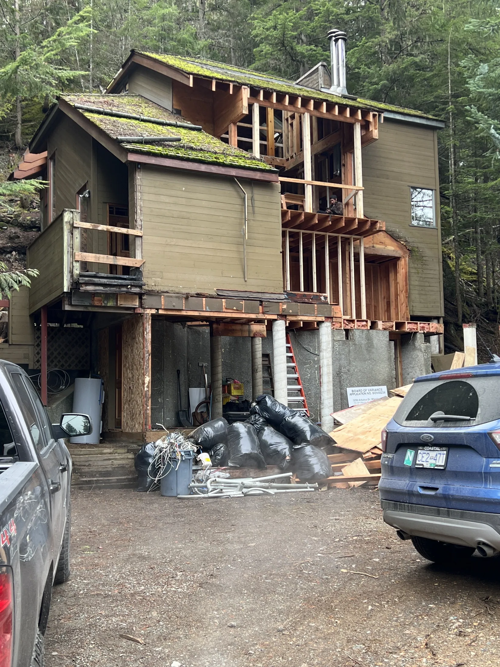 Existing cabin prior to construction, moss roof, parked vehicles and demolition debris piled in the driveway.