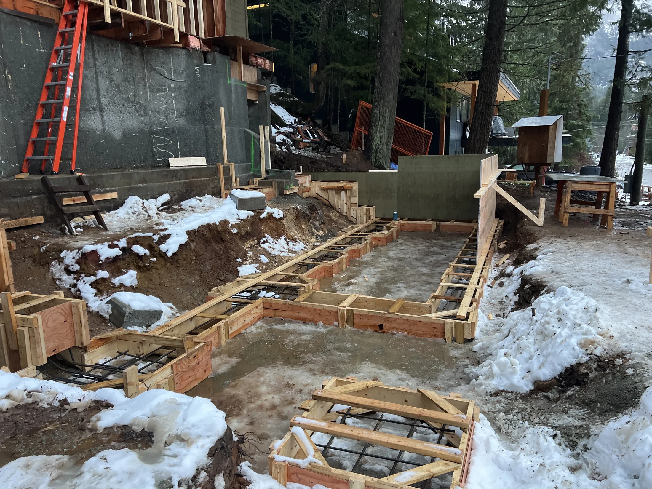 New foundation footings poured in winter snow, plywood forms and rebar before framing begins.