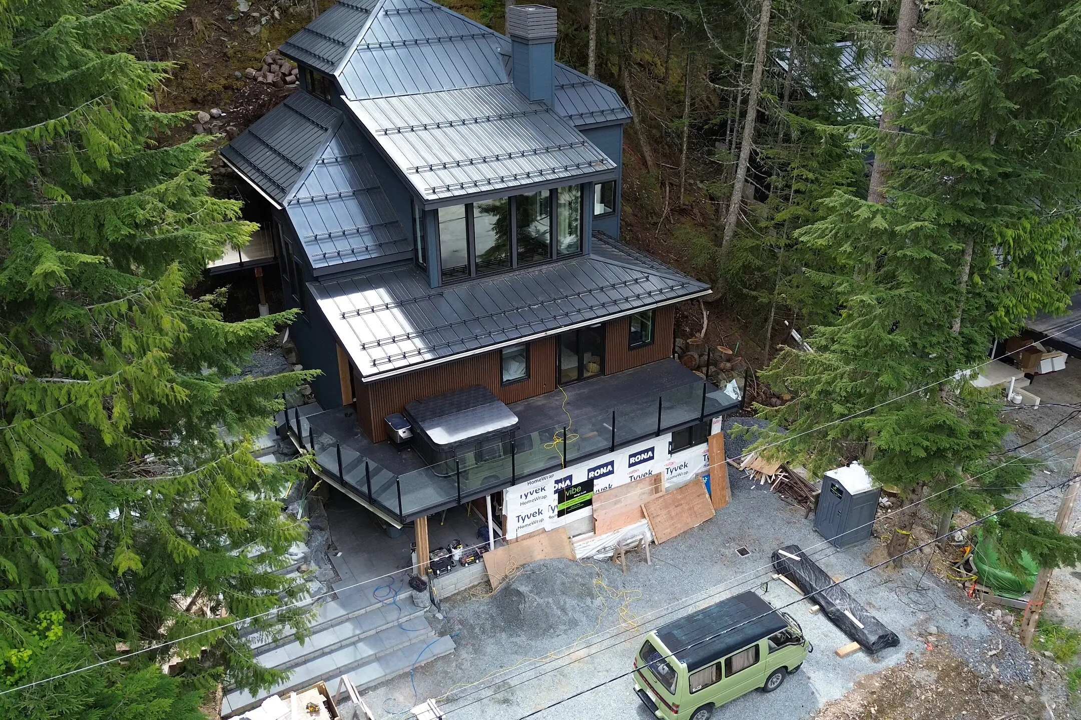 Aerial view of the home mid-construction: metal roof, Tyvek wrap, hot tub deck, dense forest surround.